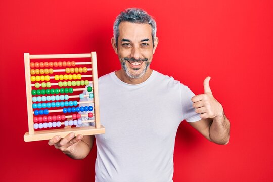 Handsome middle age man with grey hair holding traditional abacus smiling happy and positive, thumb up doing excellent and approval sign