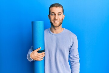 Young caucasian man holding yoga mat looking positive and happy standing and smiling with a...