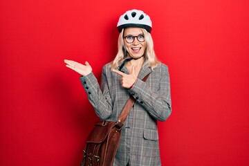 Beautiful caucasian blonde business woman wearing bike helmet amazed and smiling to the camera while presenting with hand and pointing with finger.