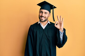Young hispanic man wearing graduation cap and ceremony robe smiling positive doing ok sign with...