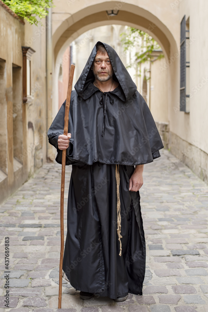 Wall mural Portrait of an elderly monk 45-50 years old with a beard and a black cassock, walking down the street with a staff against the background of a textured wall Concept: pilgrimage to holy places.