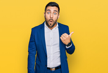 Young hispanic man wearing business jacket surprised pointing with hand finger to the side, open mouth amazed expression.