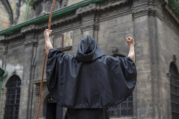 Obraz na płótnie Canvas A black-robed monk with a staff stands in front of an old, gloomy building with his staff held high.Concept: curse and mysticism, witchcraft and magic.