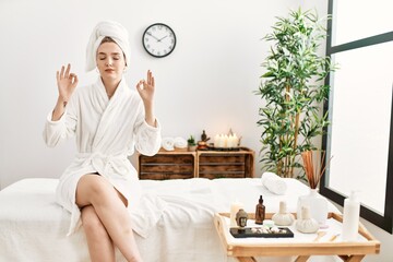 Young blonde woman wearing bathrobe at wellbeing spa relax and smiling with eyes closed doing meditation gesture with fingers. yoga concept. - obrazy, fototapety, plakaty