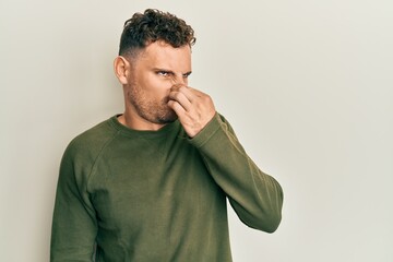 Young hispanic man wearing casual clothes smelling something stinky and disgusting, intolerable smell, holding breath with fingers on nose. bad smell