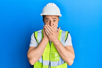 Handsome young man wearing safety helmet and reflective jacket shocked covering mouth with hands for mistake. secret concept.