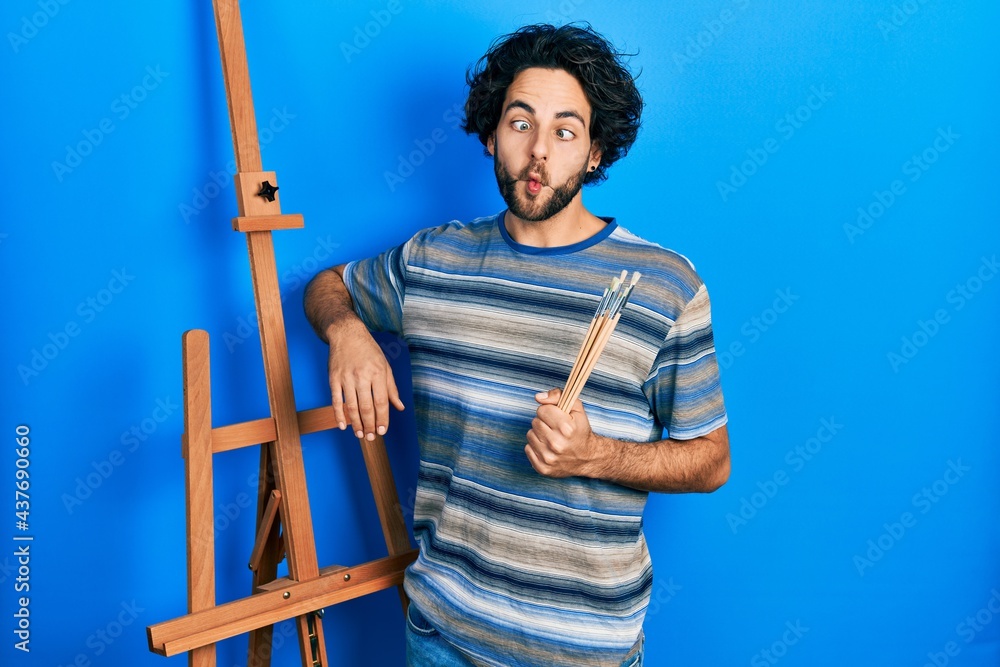 Poster Handsome hispanic man standing by empty easel stand holding brushes making fish face with mouth and squinting eyes, crazy and comical.