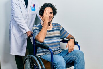 Handsome hispanic man sitting on wheelchair wearing neck collar covering one eye with hand,...