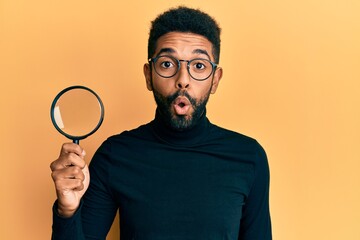 Handsome hispanic man with beard holding magnifying glass scared and amazed with open mouth for...