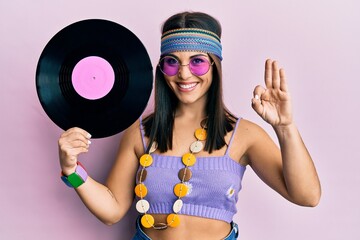 Young brunette woman wearing hippie style holding vinyl disc doing ok sign with fingers, smiling...