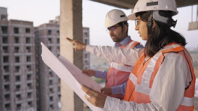 close view arc shot of young indian asian male and female civil engineers discussing a blueprint of project together standing on top of under development building near construction site