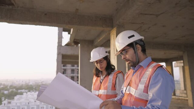 close view shot of young indian asian male and female civil engineers discussing a blueprint of project together standing on top of under development building near construction site