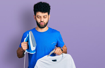 Young arab man with beard holding electric steam iron and white t shirt making fish face with mouth and squinting eyes, crazy and comical.