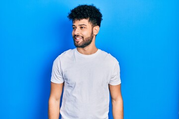 Young arab man with beard wearing casual white t shirt looking away to side with smile on face,...