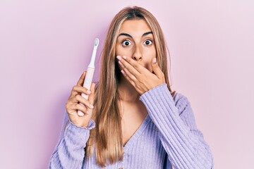 Beautiful hispanic woman holding electric toothbrush covering mouth with hand, shocked and afraid...