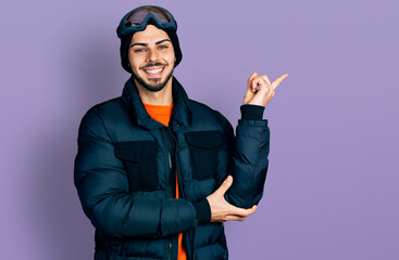 Young hispanic man with beard wearing snow wear and sky glasses with a big smile on face, pointing with hand and finger to the side looking at the camera.