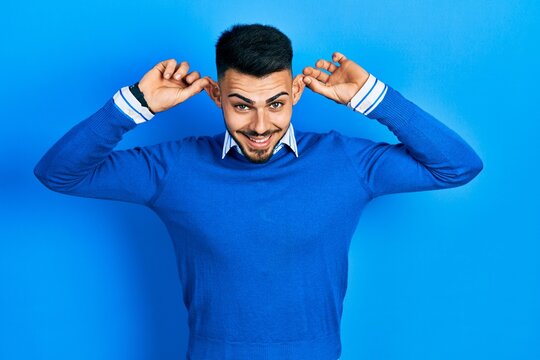 Young hispanic man with beard wearing casual blue sweater smiling pulling ears with fingers, funny gesture. audition problem
