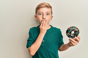 Little caucasian boy kid holding chocolate doughnut covering mouth with hand, shocked and afraid...