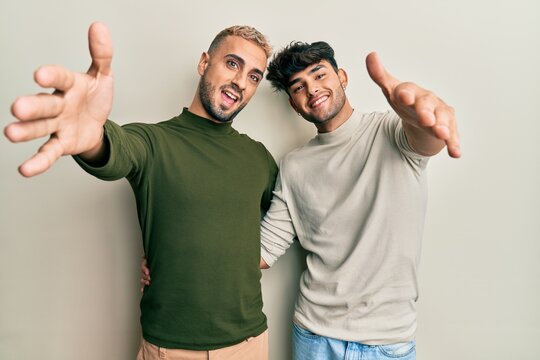 Homosexual gay couple standing together wearing casual clothes looking at the camera smiling with open arms for hug. cheerful expression embracing happiness.