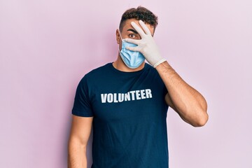 Hispanic young man wearing medical mask and volunteer t shirt peeking in shock covering face and eyes with hand, looking through fingers afraid