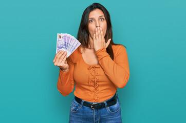 Beautiful hispanic woman holding 20 swedish krona banknotes covering mouth with hand, shocked and afraid for mistake. surprised expression