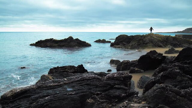 A girl tourist spreading her hands on a rocky beach cliff during sunset in 4K.  A young female exploring St Michael's Chapel Of Lammana with rocks, sand and blue water in Looe, Cornwall, England.