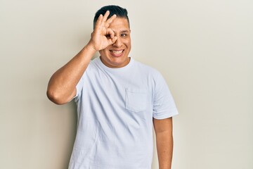 Fototapeta premium Young latin man wearing casual white t shirt doing ok gesture with hand smiling, eye looking through fingers with happy face.