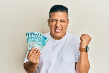 Young latin man holding 100 brazilian real banknotes screaming proud, celebrating victory and...