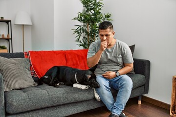 Young latin man and dog sitting on the sofa at home feeling unwell and coughing as symptom for cold...