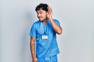 Young hispanic man wearing blue male nurse uniform smiling with hand over ear listening an hearing to rumor or gossip. deafness concept.
