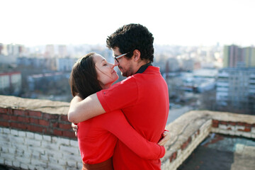 Man and caucasian woman hug each other tightly and kiss on the roof of a tall building overlooking the city. Lovers love each other and both are dressed in red.