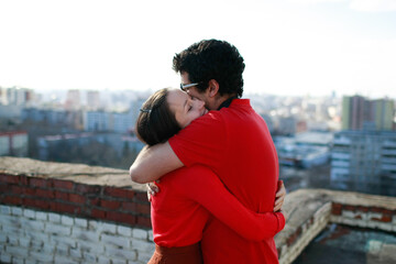 Man and caucasian woman hug each other tightly and kiss on the roof of a tall building overlooking the city. Lovers love each other and both are dressed in red.