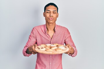 Young african american guy holding tray with cake sweets looking at the camera blowing a kiss being lovely and sexy. love expression.