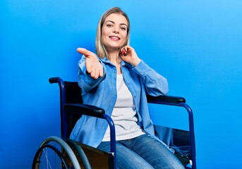 Beautiful caucasian woman sitting on wheelchair smiling friendly offering handshake as greeting and...