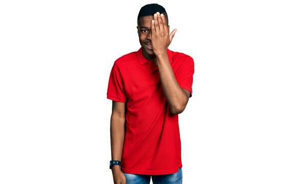 Young african american man wearing casual red t shirt covering one eye with hand, confident smile on face and surprise emotion.