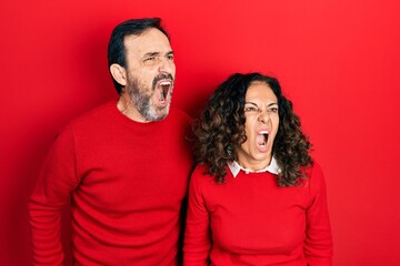 Middle age couple of hispanic woman and man hugging and standing together angry and mad screaming frustrated and furious, shouting with anger. rage and aggressive concept.