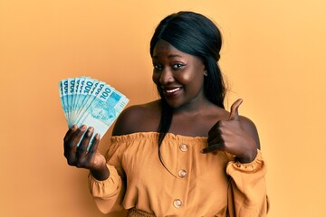 African young woman wearing doctor uniform holding 100 brazilian reals smiling happy and positive,...