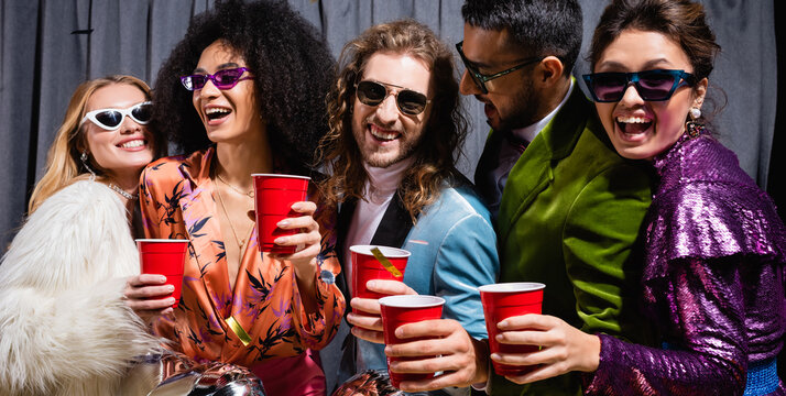 playful interracial friends in sunglasses celebrating with plastic cups on grey background, banner.