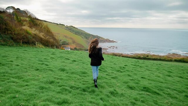 A young girl tourist running in slow motion towards Talland Bay Beach in 4K. Talland Bay, Porthallow - beautiful scenic rocky location in Cornwall, England with Holiday Houses on the seashore edge.