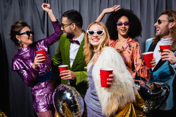 funny interracial friends in colorful clothes with plastic cups in hands dancing on grey background.