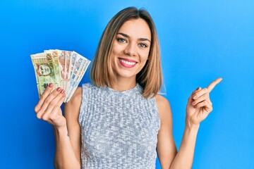 Young caucasian blonde woman holding peruvian sol banknotes smiling happy pointing with hand and finger to the side - obrazy, fototapety, plakaty