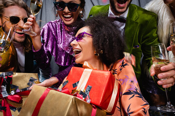 Obraz premium smiling african american woman with gift boxes sitting among interracial friends on grey background.