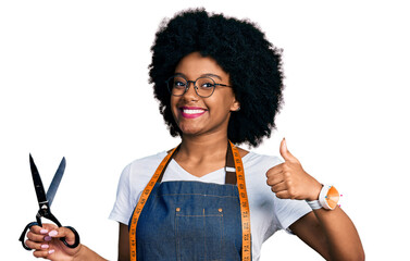 Young african american woman dressmaker designer wearing atelier apron holding scissors smiling happy and positive, thumb up doing excellent and approval sign - Powered by Adobe