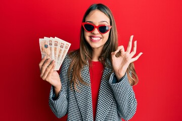 Young brunette woman holding united kingdom 10 pounds banknotes doing ok sign with fingers, smiling friendly gesturing excellent symbol