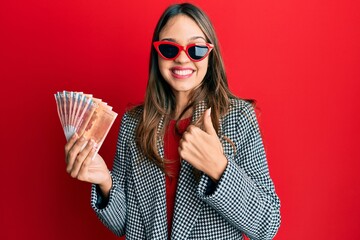 Young brunette woman holding 100 norwegian krone banknotes smiling happy and positive, thumb up...