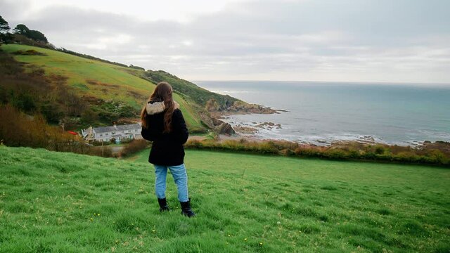 A young girl tourist watching at the Talland Bay Beach - a quiet place in 4K. Talland Bay, Porthallow - beautiful scenic rocky location in Cornwall, England with Holiday Houses on the seashore edge.