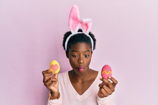 Young african american girl wearing cute easter bunny ears holding painted eggs making fish face with mouth and squinting eyes, crazy and comical.