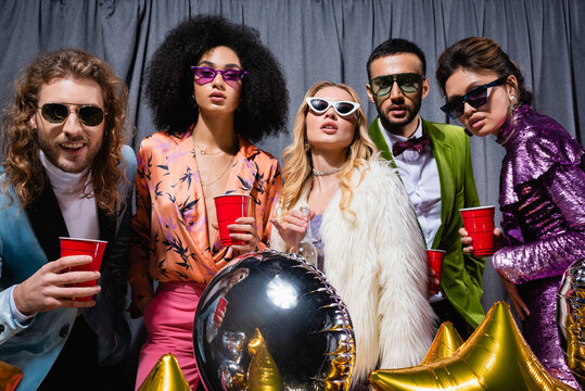 glamorous interracial friends in sunglasses standing on grey background.