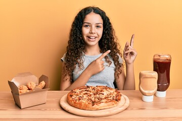 Teenager hispanic girl eating pizza and fried chicken smiling and looking at the camera pointing with two hands and fingers to the side.