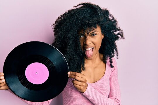 African american woman with afro hair holding vinyl disc sticking tongue out happy with funny expression.